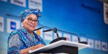 Senior woman in a blue patterned outfit and head wrap speaks at a podium during a conference.