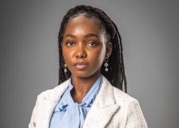 Professional headshot of a Black woman with braided hair, wearing a light blazer and pale blue blouse, against a gray studio backdrop.
