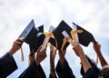 Group of graduates in navy gowns throwing their black mortarboards with gold tassels into the air against a blue sky