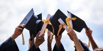 Group of graduates in navy gowns throwing their black mortarboards with gold tassels into the air against a blue sky