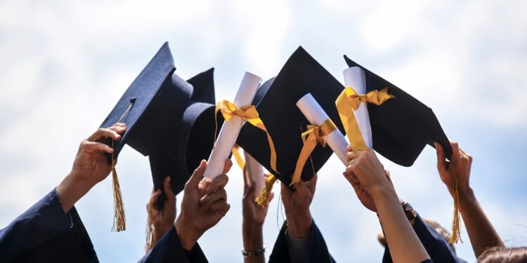 Group of graduates in navy gowns throwing their black mortarboards with gold tassels into the air against a blue sky