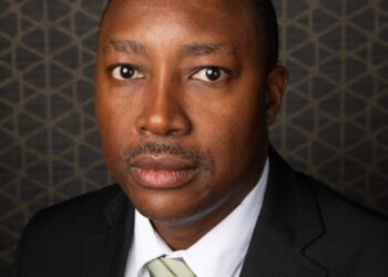 Imanuel Hawanga - The Brief | Namibia's Leading Business & Financial News Portrait of a man in a black suit and light green striped tie, looking at the camera against a geometric backdrop.