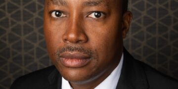 Portrait of a man in a black suit and light green striped tie, looking at the camera against a geometric backdrop.