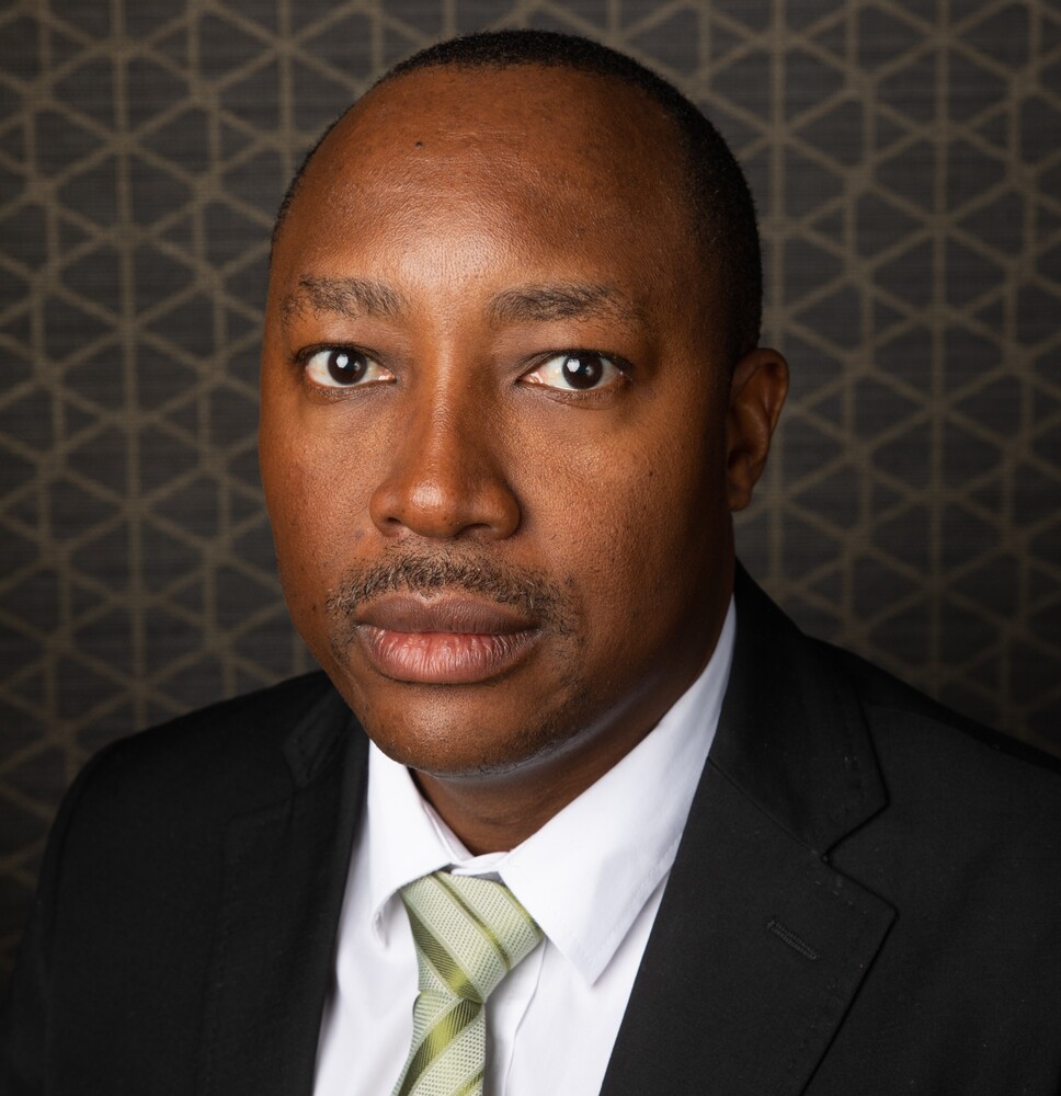 Portrait of a man in a black suit and light green striped tie, looking at the camera against a geometric backdrop.