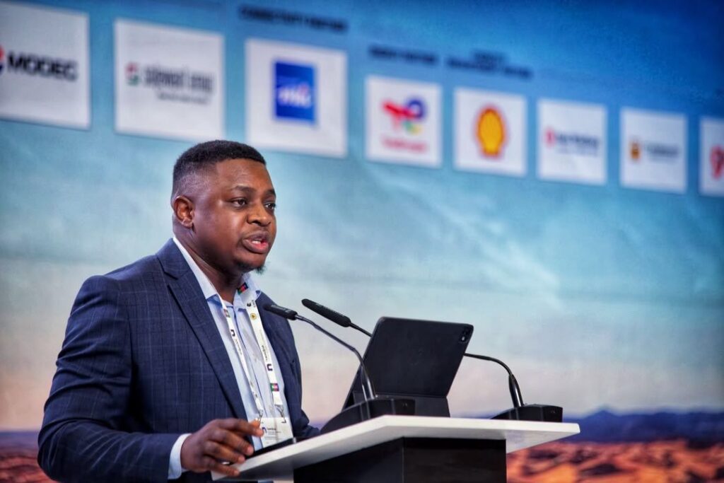 Shoki 1 - The Brief | Namibia's Leading Business & Financial News Man in a dark blue suit giving a speech at a podium with a tablet, microphones, and a sponsor-filled backdrop blurred behind him
