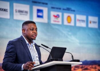 Shoki 1 - The Brief | Namibia's Leading Business & Financial News Man in a dark blue suit giving a speech at a podium with a tablet, microphones, and a sponsor-filled backdrop blurred behind him