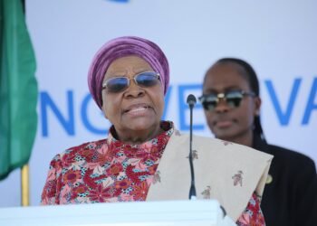 Older woman in a purple head wrap and floral dress speaking at a podium with a microphone.