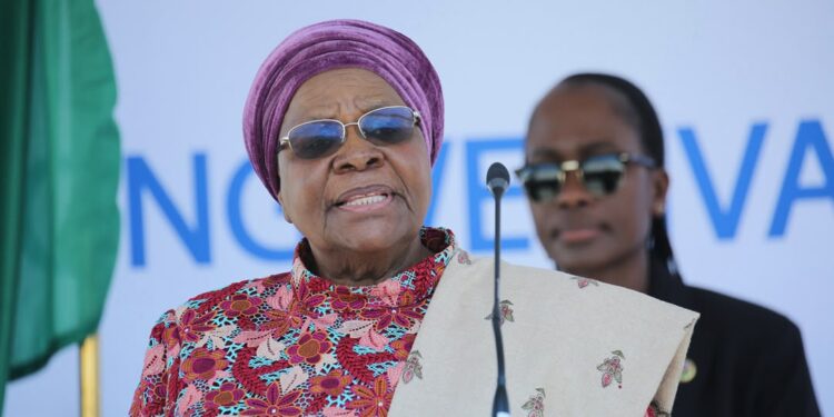 Older woman in a purple head wrap and floral dress speaking at a podium with a microphone.