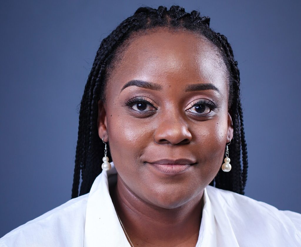 Close-up portrait of a Black woman with braided hair, pearl drop earrings, and a white shirt against a blue-gray backdrop.