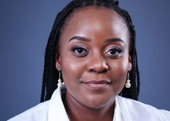 Close-up portrait of a Black woman with braided hair, pearl drop earrings, and a white shirt against a blue-gray backdrop.