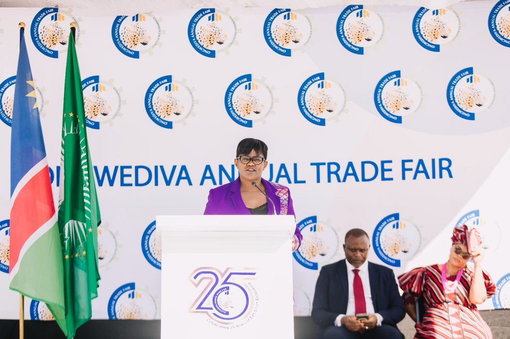 Woman in a purple blazer speaks at a podium during a trade fair stage with flags nearby and a branded backdrop reading 'Wediva Annual Trade Fair'.