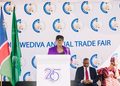 Woman in a purple blazer speaks at a podium during a trade fair stage with flags nearby and a branded backdrop reading 'Wediva Annual Trade Fair'.