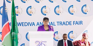 Woman in a purple blazer speaks at a podium during a trade fair stage with flags nearby and a branded backdrop reading 'Wediva Annual Trade Fair'.
