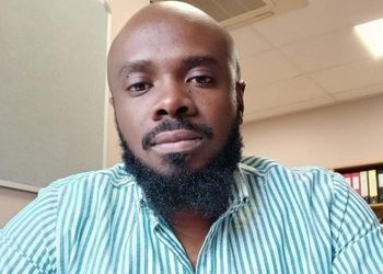 Arlindo Adriano - The Brief | Namibia's Leading Business & Financial News Close-up of a man with a full beard wearing a light teal striped shirt, seated in an office setting.