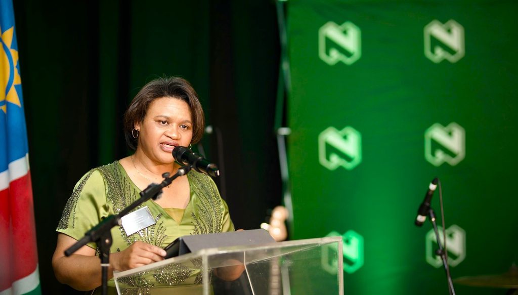 Woman in a green dress speaks at a podium with microphones, during a formal event, with a flag on the left and a green logo backdrop.