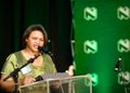 MARTHA - The Brief | Namibia's Leading Business & Financial News Woman in a green dress speaks at a podium with microphones, during a formal event, with a flag on the left and a green logo backdrop.