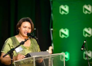 MARTHA - The Brief | Namibia's Leading Business & Financial News Woman in a green dress speaks at a podium with microphones, during a formal event, with a flag on the left and a green logo backdrop.