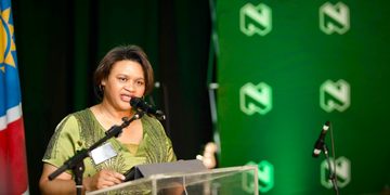 Woman in a green dress speaks at a podium with microphones, during a formal event, with a flag on the left and a green logo backdrop.
