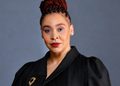 Head-and-shoulders portrait of a woman with red braided updo, black blazer, gold brooch, against a gray backdrop.