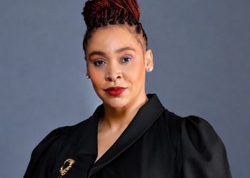 Manetti brings - The Brief | Namibia's Leading Business & Financial News Head-and-shoulders portrait of a woman with red braided updo, black blazer, gold brooch, against a gray backdrop.