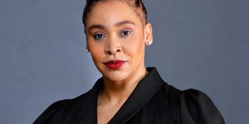 Head-and-shoulders portrait of a woman with red braided updo, black blazer, gold brooch, against a gray backdrop.