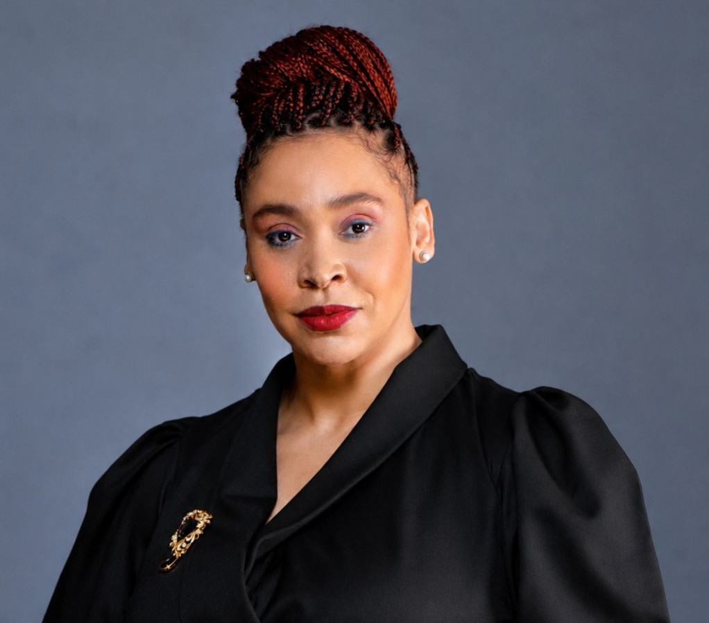 Manetti brings - The Brief | Namibia's Leading Business & Financial News Head-and-shoulders portrait of a woman with red braided updo, black blazer, gold brooch, against a gray backdrop.