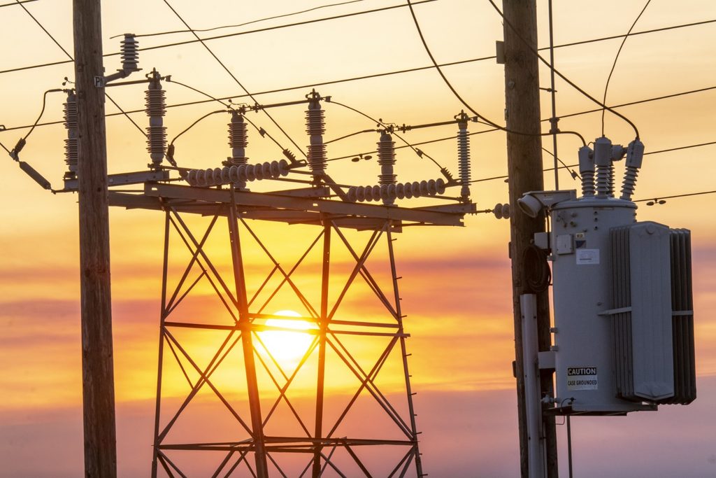 Electrical substation with transformers and hanging wires silhouetted against a sunset sky.