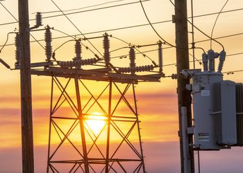 Electrical substation with transformers and hanging wires silhouetted against a sunset sky.