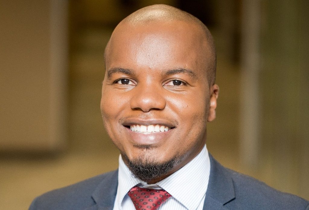 Rehabeam Ray Auala Jr - The Brief | Namibia's Leading Business & Financial News Smiling man in a navy suit with a white shirt and red tie, head-and-shoulders portrait against a blurred background.