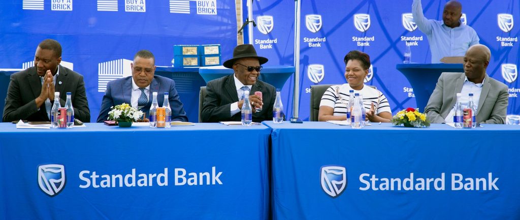 Five professionals sit at a long blue Standard Bank table during a formal event, with water bottles and small floral arrangements in front of them at a branded backdrop.