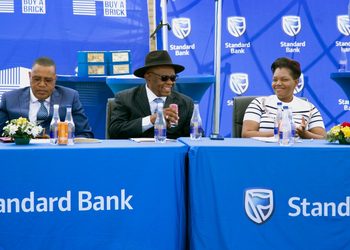 Five professionals sit at a long blue Standard Bank table during a formal event, with water bottles and small floral arrangements in front of them at a branded backdrop.