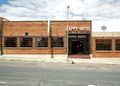 Brick hotel building with a sign reading 'KAPPS-HOTEL' on a sunny street, sidewalk in front and several wide windows on either side of the entrance.