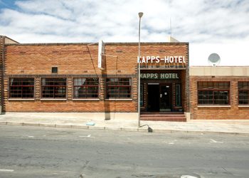 Brick hotel building with a sign reading 'KAPPS-HOTEL' on a sunny street, sidewalk in front and several wide windows on either side of the entrance.