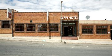 Brick hotel building with a sign reading 'KAPPS-HOTEL' on a sunny street, sidewalk in front and several wide windows on either side of the entrance.