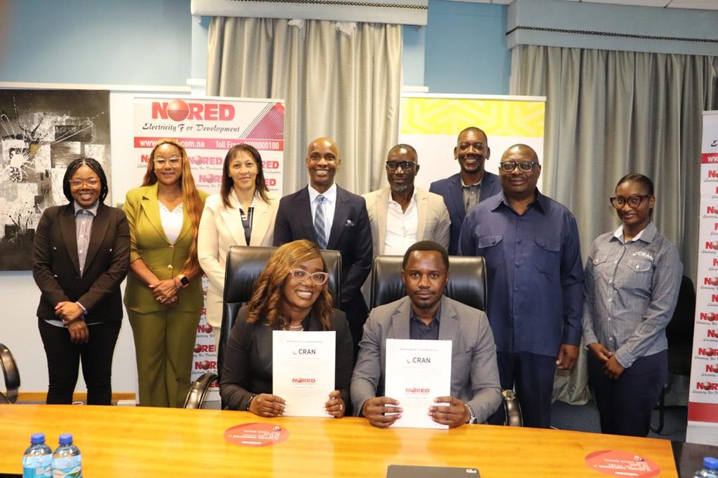 Group of ten professionals in business attire posing for a photo in an office; two people are seated at a table holding documents, while colleagues stand behind them with banners in the background.