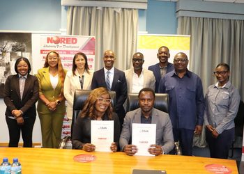 Group of ten professionals in business attire posing for a photo in an office; two people are seated at a table holding documents, while colleagues stand behind them with banners in the background.