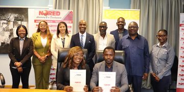 Group of ten professionals in business attire posing for a photo in an office; two people are seated at a table holding documents, while colleagues stand behind them with banners in the background.