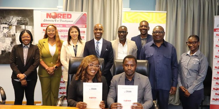 Group of ten professionals in business attire posing for a photo in an office; two people are seated at a table holding documents, while colleagues stand behind them with banners in the background.
