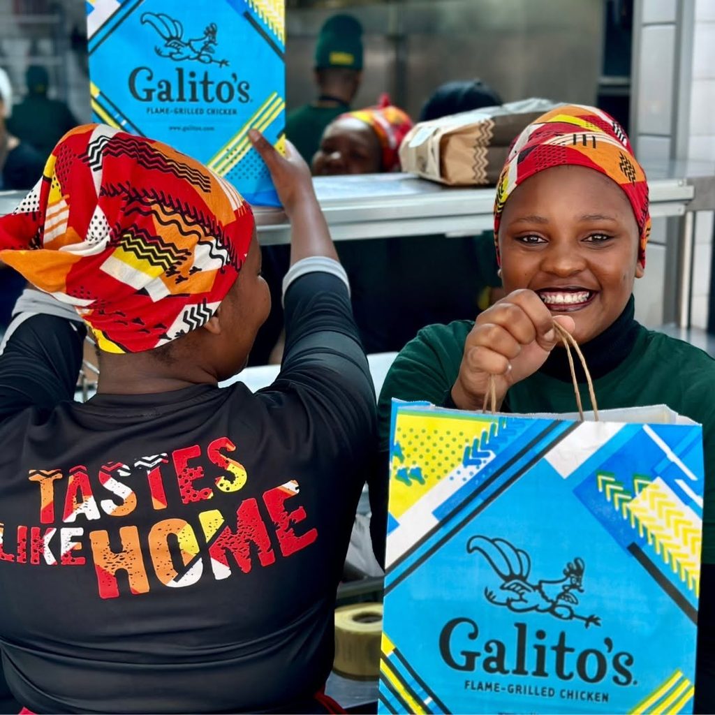 Two women wearing colorful headwraps smile as they hold bright Galito's chicken bags, with one wearing a shirt that reads 'TASTES LIKE HOME.'