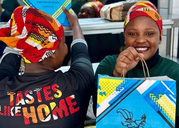 Two women wearing colorful headwraps smile as they hold bright Galito's chicken bags, with one wearing a shirt that reads 'TASTES LIKE HOME.'