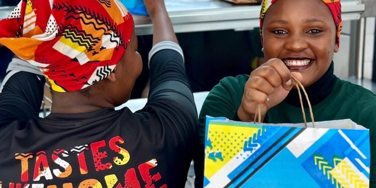 Two women wearing colorful headwraps smile as they hold bright Galito's chicken bags, with one wearing a shirt that reads 'TASTES LIKE HOME.'