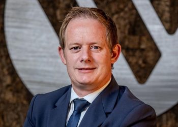 Man in a navy suit and blue tie smiling, with a textured backdrop behind him (professional portrait).**