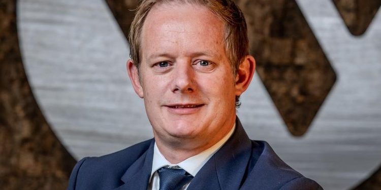 Man in a navy suit and blue tie smiling, with a textured backdrop behind him (professional portrait).**
