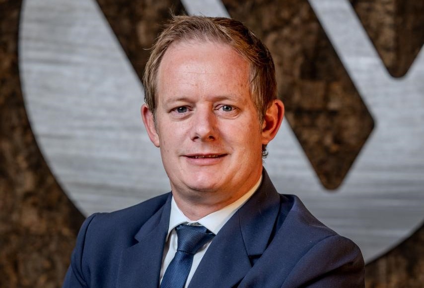 Man in a navy suit and blue tie smiling, with a textured backdrop behind him (professional portrait).**
