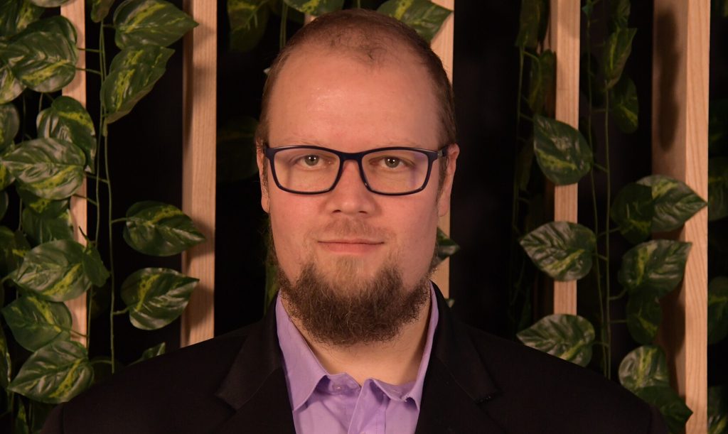 Head-and-shoulders portrait of a man with glasses and a beard, wearing a purple shirt and black blazer, in front of a plant-filled backdrop.