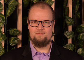 Head-and-shoulders portrait of a man with glasses and a beard, wearing a purple shirt and black blazer, in front of a plant-filled backdrop.