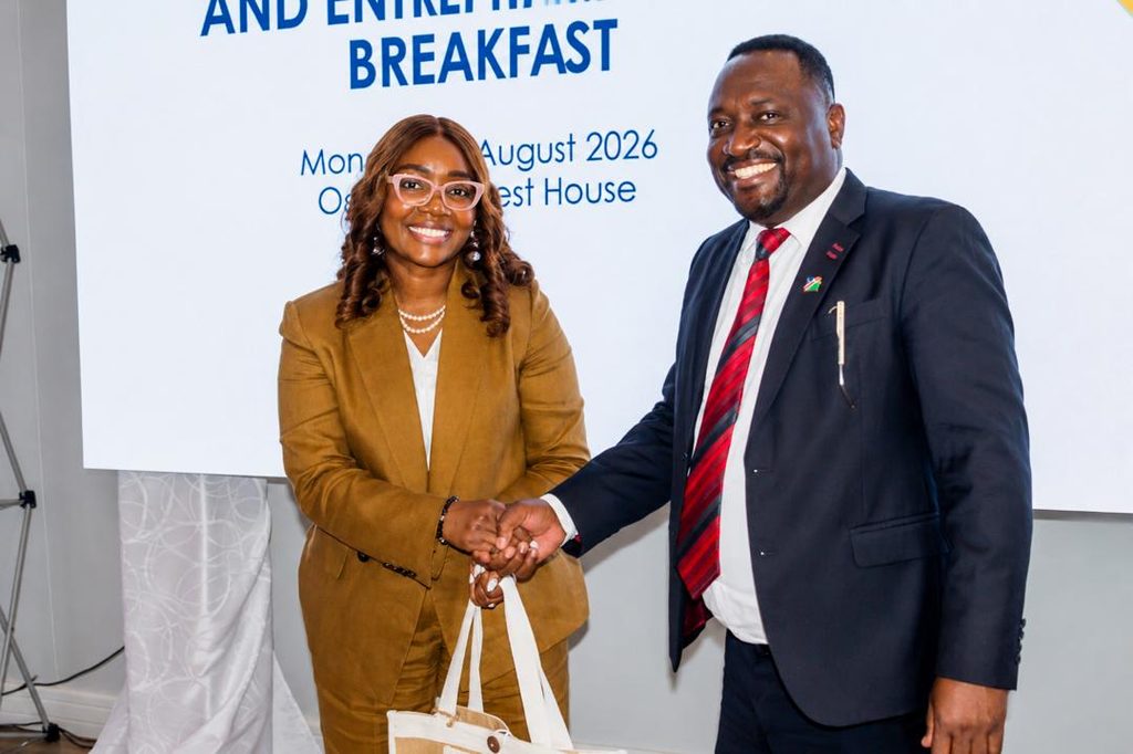 Two professionals shake hands and smile at a formal event, with a banner reading 'BREAKFAST' behind them. The woman wears a tan suit; the man wears a navy suit and red tie.