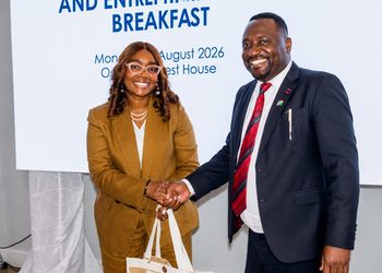 Two professionals shake hands and smile at a formal event, with a banner reading 'BREAKFAST' behind them. The woman wears a tan suit; the man wears a navy suit and red tie.