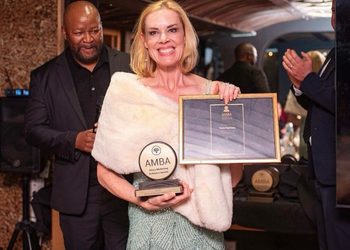 Woman in a white fur stole holds an AMBA trophy and a framed certificate at an awards ceremony, with a man in a suit behind her.