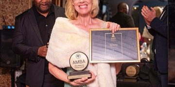 Woman in a white fur stole holds an AMBA trophy and a framed certificate at an awards ceremony, with a man in a suit behind her.